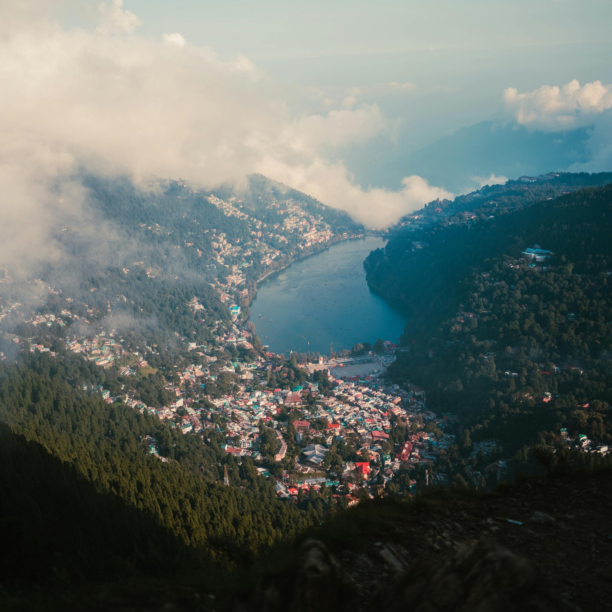 A breathtaking aerial view of Nainital Lake surrounded by mountains and clouds under a clear sky.